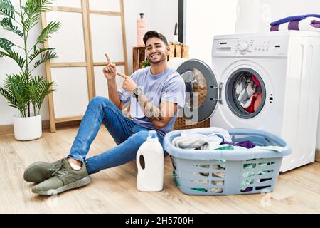 Jeune hispanique mettant le linge sale dans le lave-linge souriant et regardant la caméra pointant avec deux mains et les doigts sur le côté. Banque D'Images