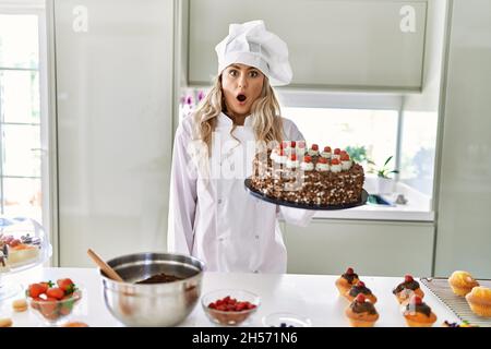 Jeune chef de pâtisserie caucasienne femme cuisant des pâtisseries et des gâteaux à la cuisine effrayée et stupéfaite avec la bouche ouverte pour la surprise, le visage incrédulé Banque D'Images