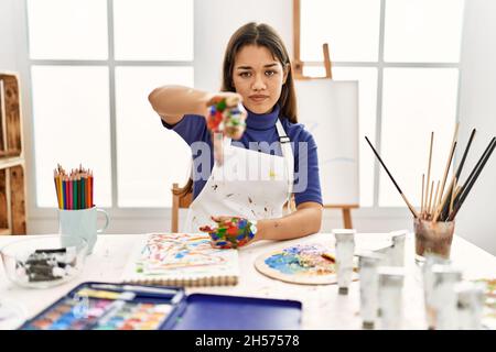 Jeune femme brune au studio d'art avec des mains peintes regardant malheureux et en colère montrant le rejet et négatif avec les pouces vers le bas geste. Mauvaise expressio Banque D'Images