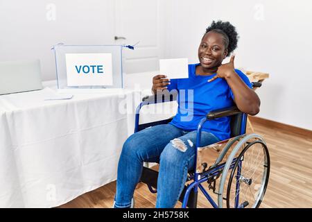 Jeune femme africaine assise sur un fauteuil roulant en train de voter en plaçant une enveloppe dans l'urne en souriant en faisant un geste de téléphone avec la main et les doigts comme en parlant sur la t Banque D'Images