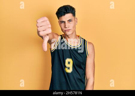 Jeune homme hispanique portant un uniforme de basket-ball à l'air malheureux et en colère montrant le rejet et négatif avec le pouce vers le bas geste. Mauvaise expression. Banque D'Images