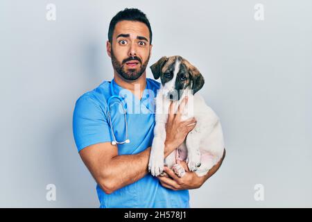 Beau vétérinaire hispanique avec la barbe de vérifier la santé de chien dans le visage de choc, regardant sceptique et sarcastique, surpris par la bouche ouverte Banque D'Images