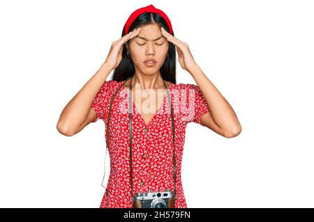 Belle jeune femme asiatique portant robe et appareil photo vintage souffrant de maux de tête désespérés et stressés parce que la douleur et la migraine. Mains sur la tête. Banque D'Images