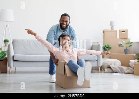 Satisfait riant jeune mari afro-américain poussant boîte avec une femme excitée sur le nouvel appartement dans le salon Banque D'Images