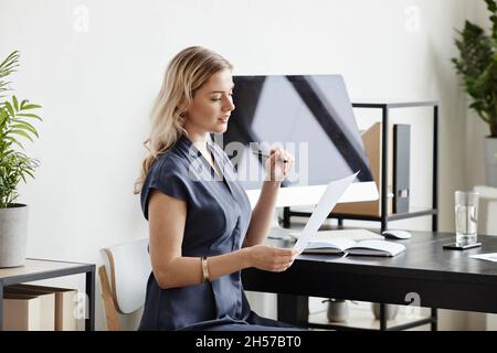 Portrait de femme d'affaires blonde élégante avec vue latérale, lisant des documents tout en travaillant au bureau, dans un espace de travail Banque D'Images