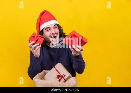 Un jeune homme dans un chapeau de Père Noël montre des cadeaux de Noël heureux.Fond jaune pour un portrait de studio.Joyeux Noël concept. Banque D'Images