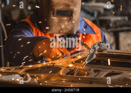 Le soudeur industriel dans un masque de protection effectue des travaux de soudage avec des étincelles et répare la porte de la voiture dans l'atelier de l'usine. Banque D'Images