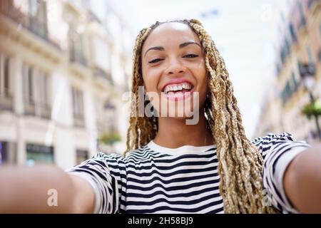 Femme noire avec des tresses afro prenant un selfie souriant dans une rue urbaine avec un smartphone. Banque D'Images