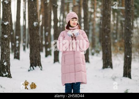 Portrait d'hiver extérieur d'une jolie jeune femme portant un manteau rose et un élégant bonnet tricoté et une écharpe, debout dans une belle forêt enneigée devant des pins et profitant de la marche par temps froid Banque D'Images