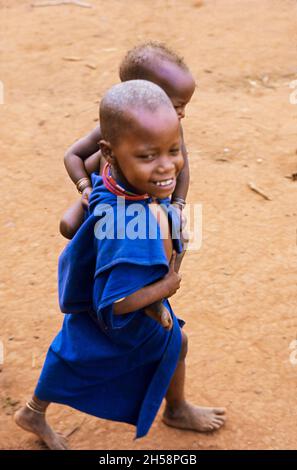 Afrique, Tanzanie, 1976.Le village d'Ujamaa Maasai les gens vivent encore dans leurs maisons traditionnelles.Un jeune enfant transportant un bébé. Banque D'Images