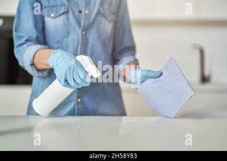 Gros plan des mains des femmes dans des gants en caoutchouc à l'aide d'un détergent en aérosol et d'un chiffon lors du nettoyage de la surface de la cuisine Banque D'Images