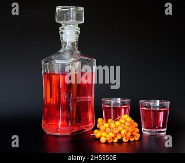 Liqueur maison sur frêne de montagne, alcool fort dans une bouteille de verre et deux verres sur fond noir, à côté d'un bouquet de baies mûres.Arrière-plan, Banque D'Images