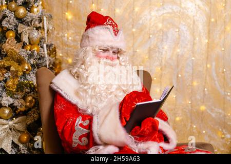 Le Père noël est assis sur une chaise, contre le fond des écrits de l'arbre de noël dans le carnet.Regarde la liste des cadeaux de noël pour les enfants.Réalisation des désirs à la veille du nouvel an. Banque D'Images