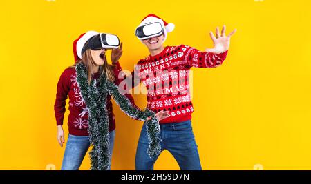 Jeune joueur de couple latin en réalité virtuelle casque gadget portant chapeau de Santa et chandail de Noël sur fond jaune.Noël et hiver Banque D'Images