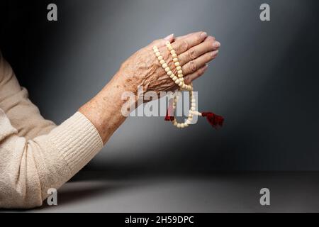 Les mains froissées d'une femme âgée se replient dans la prière et tiennent un rosaire.Copier l'espace.Arrière-plan gris.Le concept de religion. Banque D'Images