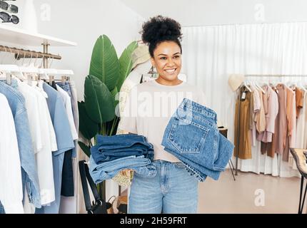 Une vendeuse gaie debout dans un magasin de vêtements avec deux piles de jeans Banque D'Images