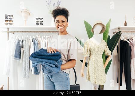 Une vendeuse souriante se tenant dans un petit magasin local avec une pile de jeans et regardant loin Banque D'Images