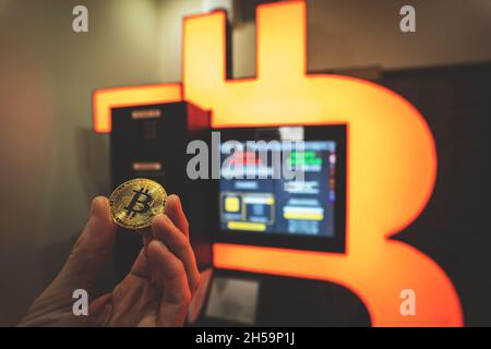 Pièce d'or Bitcoin tenue à la main à côté de Bitcoin ATM, machine d'échange de crypto-monnaie orange qui permet d'acheter et de vendre la crypto de BTC.Image teintée avec mise au point sélective. Banque D'Images