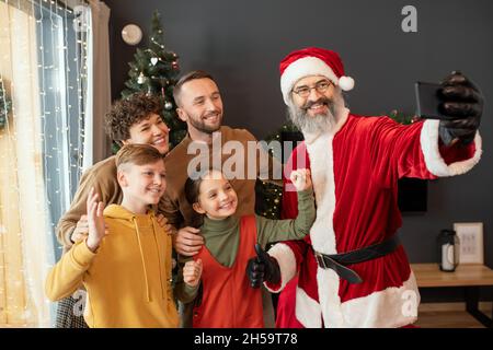 Sourire le Père Noël en costume rouge prenant le selfie avec la famille heureuse sur le smartphone contre l'arbre de Noël Banque D'Images