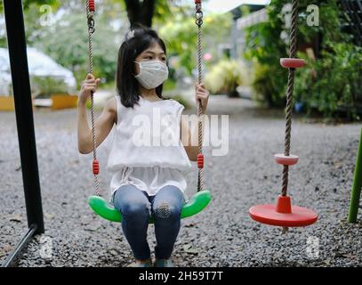 Une jeune fille asiatique mignonne avec un masque blanc joue seule sur une balançoire dans un terrain de jeu pendant la pandémie Covid-19. Banque D'Images