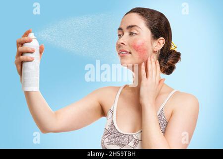 Une jeune femme du Caucase avec la rosacée sur ses joues vaporise de l'eau thermique sur son visage, obtenant le soulagement et la fraîcheur.Arrière-plan bleu.Concept de cosmétique Banque D'Images