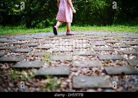 Une vue arrière moitié inférieure du corps photo d'une petite fille en robe rouge et blanche marchant sur un pavé de pierre et de gravier à l'intérieur d'un jardin avec des buissons verts en t Banque D'Images