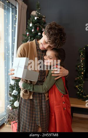 Bonne mère aux cheveux bouclés dans une robe tenant une boîte cadeau ouverte et embrassant l'enfant tout en la remerciant pour son cadeau de Noël Banque D'Images