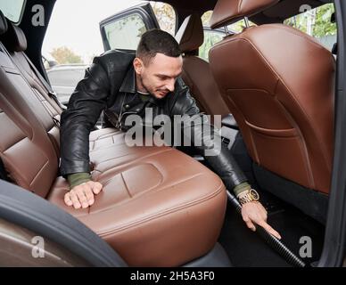 Vue de l'intérieur d'une voiture sur un jeune homme lavant une voiture sur le poste de lavage de voiture extérieur.Un travailleur de qualité nettoyant l'intérieur de l'automobile à l'aide d'un aspirateur. Banque D'Images