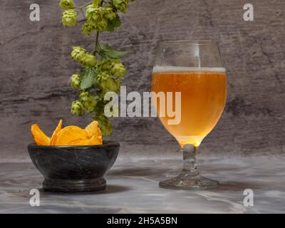 Verre de bière avec une branche de houblon et un bol avec des chips de pomme de terre sur fond gris. Banque D'Images
