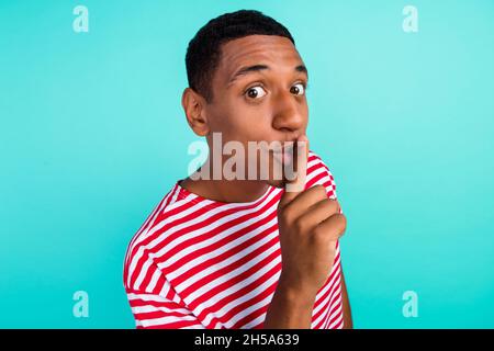 Photo de sérieux beau jeune homme porter t-shirt rayé lèvres doigt demandant garder le silence isolé teal couleur fond. Banque D'Images