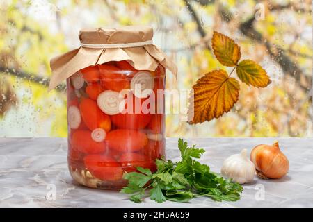 Tomates conservées dans un pot en verre, feuilles de persil, garlics, un oignon sur le windowsiil.Fenêtre avec gouttes d'eau et feuilles d'automne sur le fond. Banque D'Images