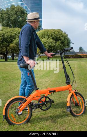 Un homme d'âge moyen se tient avec un vélo sur la pelouse d'un parc de la ville, sur le fond d'un immeuble de bureaux.Style de vie actif et environnement frie Banque D'Images