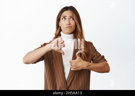 Indécis femme d'affaires d'âge moyen pesant la décision, montrant comme un geste déplaît, pouces vers le haut et pouces vers le bas, regardant vers le haut et pensant, blanc Banque D'Images