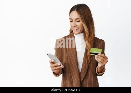 Femme d'affaires élégante utilisant une carte de crédit et un téléphone portable pour commander en ligne, payer avec un smartphone et sourire, debout sur fond blanc Banque D'Images