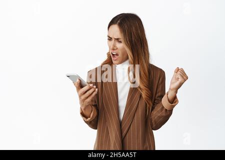 Une femme d'âge moyen en colère regardant un téléphone portable, se plaignant d'un téléphone portable, se tenant debout sur un fond blanc Banque D'Images