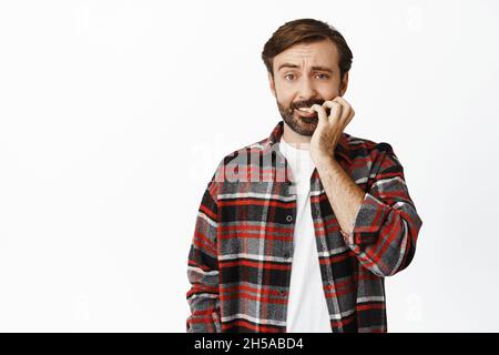 Portrait d'un homme barbu effrayé piquant ses ongles, regardant anxieux, regardant nerveusement quelque chose, debout sur fond blanc Banque D'Images