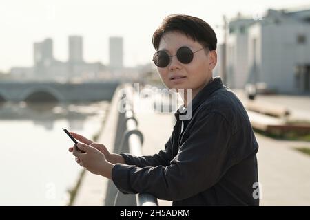 Jeune femme d'affaires chinoise dans des lunettes de soleil debout au bord de la rivière et défilant dans le smartphone Banque D'Images