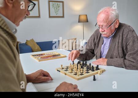 Homme senior qui fait chéquier tout en jouant aux échecs avec son ami à la table Banque D'Images