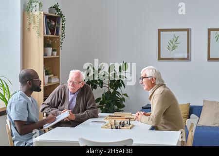 Infirmière masculine donnant des recommandations aux hommes âgés pendant qu'ils sont assis à la table pendant le jeu d'échecs dans la maison de repos Banque D'Images
