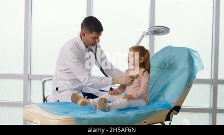 Un enfant visite le cabinet du médecin.Le docteur écoute les poumons de l'enfant avec un stéthoscope, parle avec Caringly la petite fille. Banque D'Images