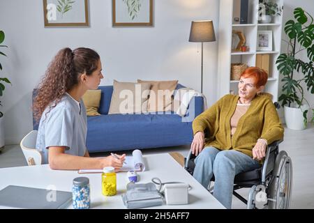 Femme sénior assise en fauteuil roulant et discutant de son traitement avec le médecin pendant qu'elle remplit sa carte médicale Banque D'Images