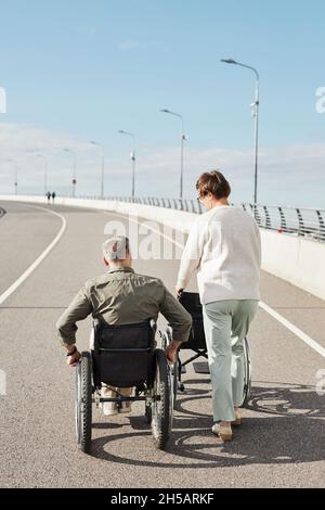 Vue arrière verticale portrait d'un couple mature avec un homme en fauteuil roulant marchant sur la route au pont, espace de copie Banque D'Images