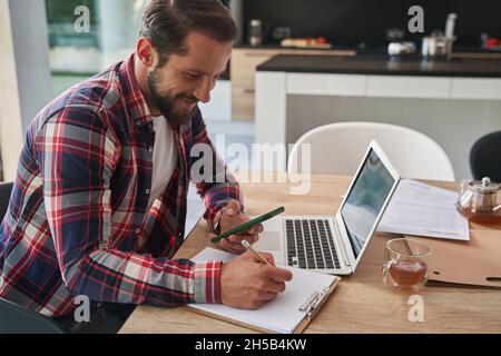 Homme élégant barbu dans une chemise écrivant des notes tout en tenant le téléphone portable dans la pièce à l'intérieur Banque D'Images