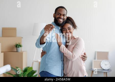 Une jeune famille affectueuse afro-américaine souriante s'embrasse dans une nouvelle maison parmi des boîtes et montre les clés de la maison Banque D'Images