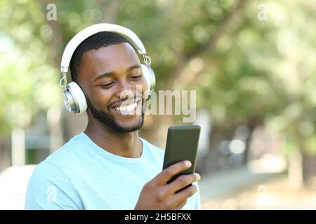 Homme heureux avec peau noire écoutant de la musique avec des écouteurs et un smartphone marchant dans un parc Banque D'Images