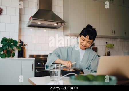 Femme souriante ouvrant le récipient à nourriture à la table dans la cuisine Banque D'Images