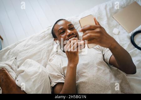 Homme souriant d'un âge adulte moyen utilisant un téléphone portable tout en étant allongé sur le lit Banque D'Images