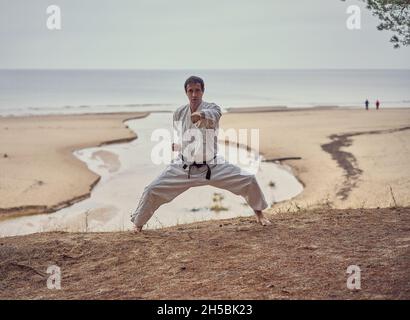 Carie dans un vieux kimono et ceinture noire punch d'entraînement dans le stand kibadachi à la mer.Concept des arts martiaux.La rivière coule dans la mer à l'arrière-plan. Banque D'Images