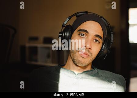 Jeune homme millénaire à l'œil voilé de compétition mixte, qui écoute de la musique sur un casque circum-aural et regarde l'appareil photo.Taille basse avec casque et bonnet au soleil Banque D'Images