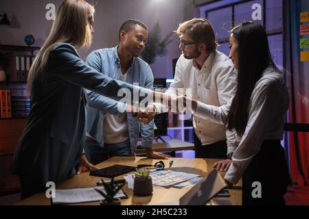 Équipe de testeurs multiculturels de logiciels qui se secouent les mains tout en se tenant près de la table avec divers gadgets et papiers.Rencontre avec un client.Une coopération réussie. Banque D'Images